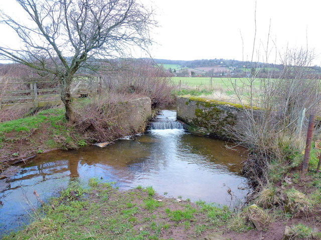 A low stone weir on a small brook spilling into a still pool, with the Golden Valley and wooded hills beyond.